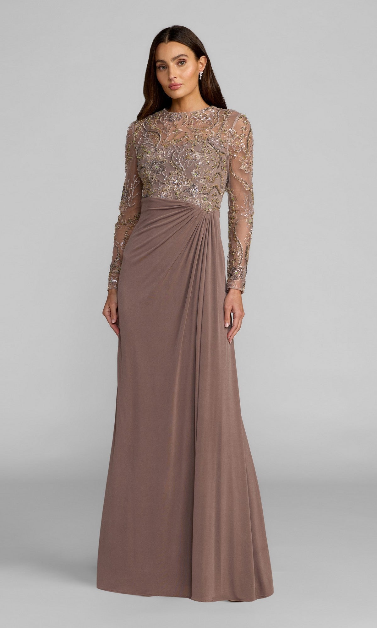 A woman wearing the Mac Duggal 69121 long formal dress, which features a sequin-embroidered bodice and sheer sleeves, stands against a plain gray background. This stunning golden brown gown is adorned with intricate beaded detailing for an elegant touch.